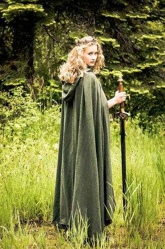 Teen or young adult girl wearing a crown and green cloak holding a long sword and looking over her shoulder against a lush forest background. Soft fantasy look.