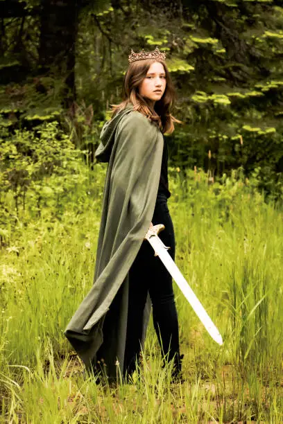 Teen or young adult girl wearing a crown and green cloak swinging a long sword and looking over her shoulder against a lush forest background. Soft fantasy look