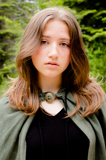 Teen or young adult girl wearing a green cloak looking at the camera against a lush forest background. Soft fantasy look