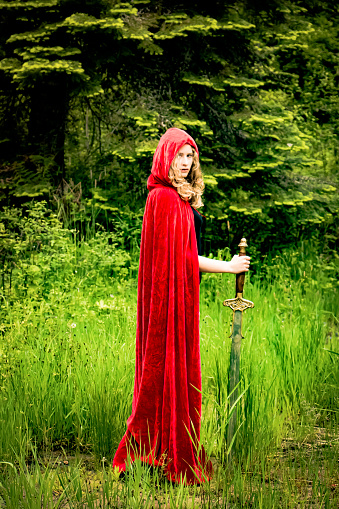 Teen or young adult girl wearing a crown and red cloak holding a long sword and looking over her shoulder against a lush forest background. Soft fantasy look.