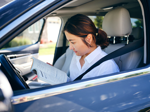 An Asian Korean woman studying from a textbook while waiting in a car.