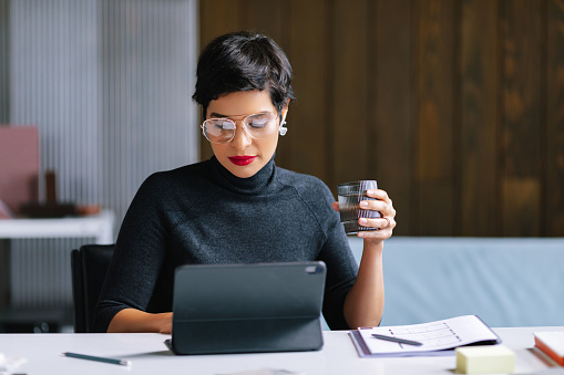 Latin businesswoman sitting at a working desk, holding a glass of water, typing on her notepad.