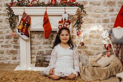 Cute girl in white dress sitting on floor by the fireplace and looking at camera during Christmas and New Year holidays at home