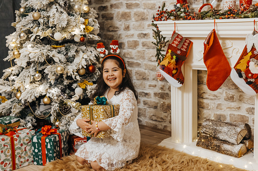 Happy girl in white dress celebrating Christmas and New Year holidays at home, sitting on floor and holding Christmas present
