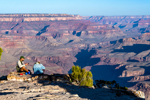A young couple is sitting on the south rim of the Grand Canyon. The image shows them having breakfast and looking at a dramatic canyon view from edge of a precipice in early morning light.\nGrand Canyon National Park\n05/17/22
