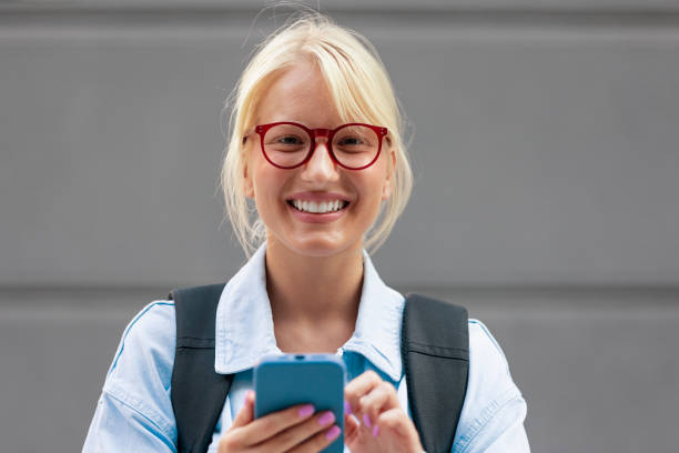 可愛い女子生徒がスマホを握りしめ、カメラに向かって微笑んでいるクローズアップ写真。 - real people blond hair denim youth culture ストックフォトと画像