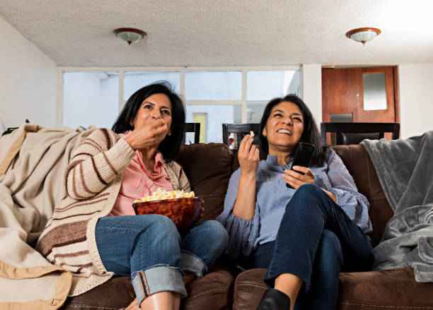 istock two women sitting on a couch, eating popcorn and laughing while watching television 1417451981