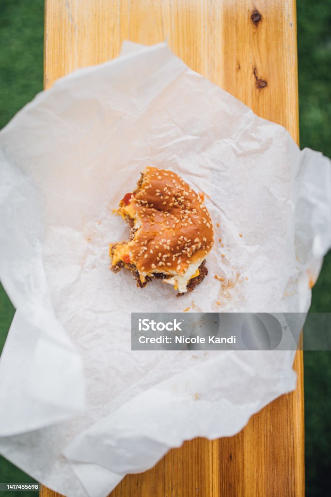 high angle view of half eaten cheeseburger on paper eating a fast food cheeseburger with a sesame bun, outdoors on bench Burger Stock Photo