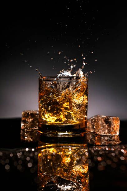 bourbon whiskey or rum splashing out of a glass with ice 
cubes on black reflection - action alcohol alcoholism bar imagens e fotografias de stock