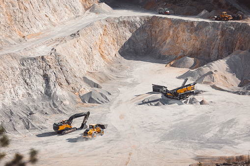 Heavy machinery in the quarry, excavator and small dump track working in the quarry. Piles of gravel all around, machines moving the gravel. Dusty roads in the quarry.