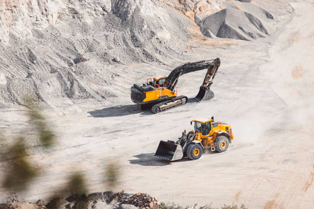 Heavy machinery in the quarry, excavator and small dump track working in the quarry. Piles of gravel all around, machines moving the gravel. Dusty roads in the quarry.