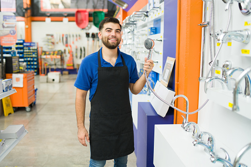 Smiling young man and hardware store employee wearing a blue apron showing a shower head to a customer shopping
