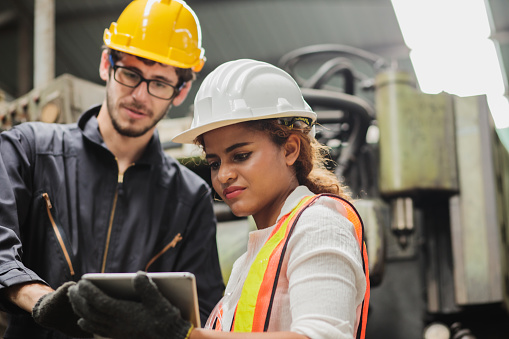 Female industrial engineer wearing a white helmet while standing in a heavy industrial factory behind she talking with workers, Various metal parts of the project