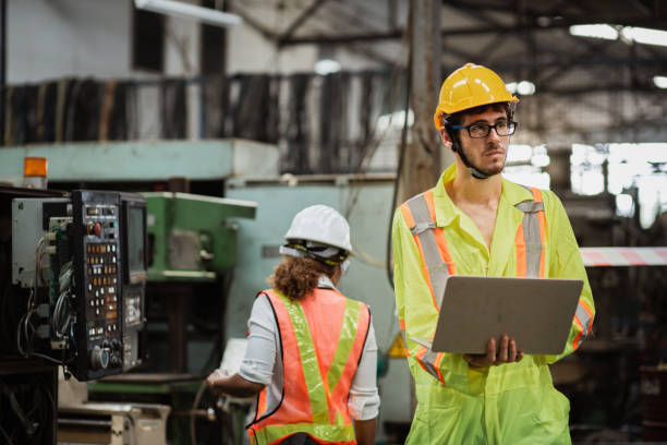 istock Men industrial engineer in uniform and wearing a Yellow helmet while standing in a heavy industrial factory behind he looking New Project while Using Laptop. 1417486776