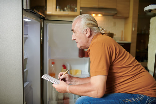 Senior man checking his fridge and making shopping list before going to grocery store