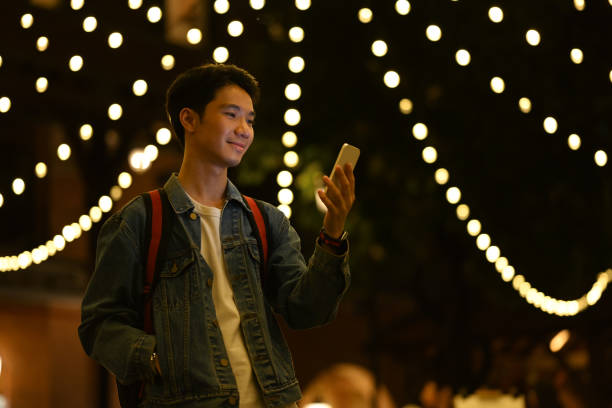 Smiling young hipster man using smart phone while walking at evening city streets.