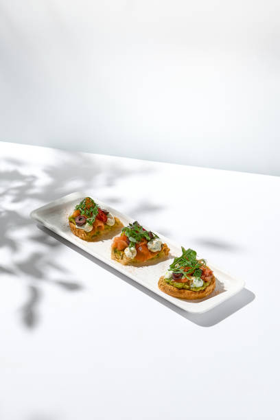 istock Aesthetic composition with salmon bruschetta on white background with shadows from flowers. Italian bruschetta with salmon, avocado, cheese and olives on fine dining in summer. Elegant menu concept. 1417559825