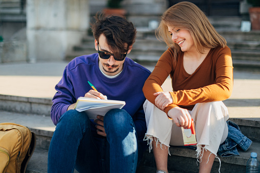 A smiling woman and a focused man sitting on the stairs. He writes down important information about the exam