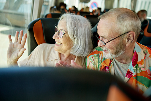 Head and shoulders view of cheerful man and woman in 60s and 70s looking out panoramic bus window and gesturing goodbye.