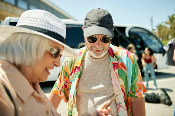 Senior couple checking smart phone before boarding coach bus Close view of casually dressed man and woman wearing hats and sunglasses as they stand in late spring sunshine using portable device while waiting to depart. coach bus stock pictures, royalty-free photos & images