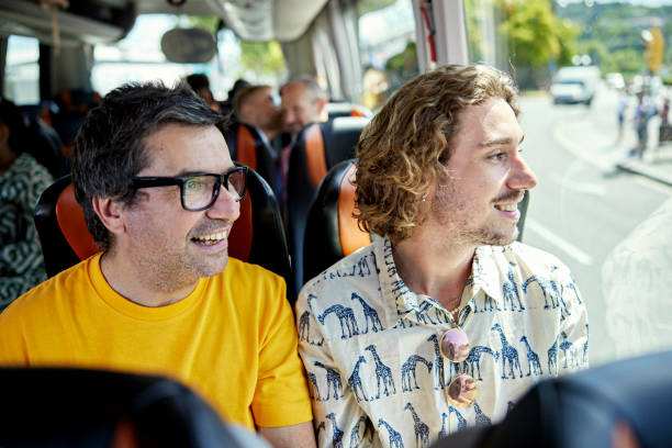 Casually dressed man in mid 50s and adult son in early 20s enjoying panoramic window and scenery while onboard bus.