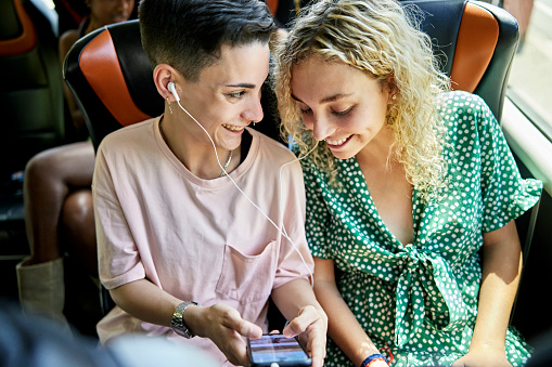 Waist-up view of smiling women in late teens and 20s sitting side by side onboard deluxe motor coach and enjoying smart phone entertainment.