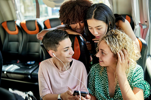 Waist-up view of casually dressed female friends in late teens and 20s gathered around smart phone while traveling by bus to their destination.