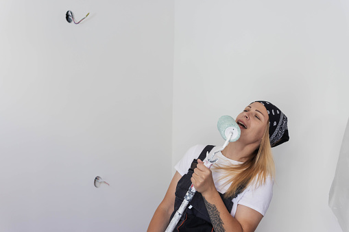 cheerful girl in a bandana and work overalls,stands posing at the camera with roller for painting walls and sings songs cheerfully in the roller standing facing the camera cheerful. High quality photo