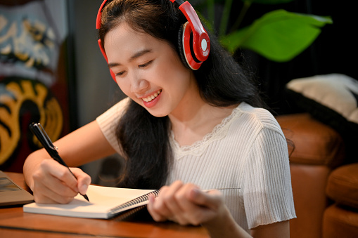 Attractive beautiful young Asian female listening to music on her headphones and writing her diary in the living room.