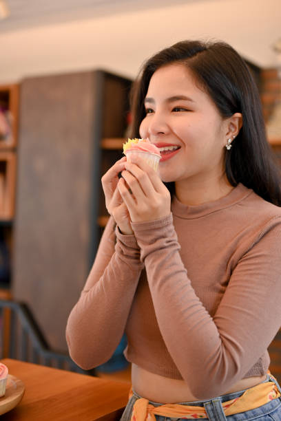 giovane bella donna asiatica che mangia cupcakes in salotto, piena di gioia e felice - thai cuisine asian cuisine vertical close up foto e immagini stock