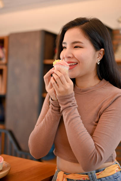 jovem linda mulher asiática comendo cupcakes na sala de estar, cheia de alegria e feliz - thai cuisine asian cuisine vertical close up - fotografias e filmes do acervo