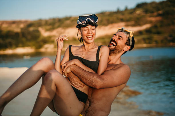 Fun couple at the beach Spontaneous image of a young adult, attractive couple enjoying their time at the beach, being playful after a swim, while in their swimsuits. Both nature lovers, adventure seekers, having a snorkeling masks on their heads. Smiling, radiating positivity. A handsome guy is hugging and spinning his darling brunet. Both radiating fun, love enjoyment healthy slim fit in bikini stock pictures, royalty-free photos & images
