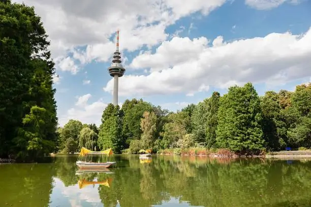 The Telecommunications Tower in the Luisenpark park in Mannheim, Germany, with a pond and lush green trees in the background
