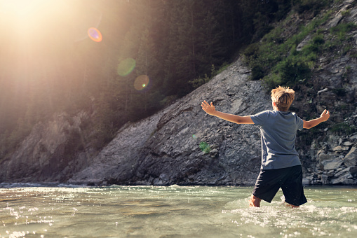 Teenage boy is hiking in the Alps - Vorarlberg, Austria. The boy is standing in waters of river Lech and enjoying the sun.\nCanon R5