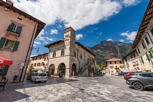 Venzone, Italy - August 11, 2022: Ancient town hall building and square (Piazza Municipio) in the small village of Venzone, partially destroyed by the 1976 earthquake and rebuilt between 1979 and 1984. Udine province, Friuli-Venezia Giulia, Italy, Europe. A group of locals and tourists stroll through the ancient square on a sunny summer day.