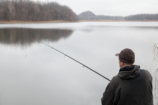 A man in a jacket catches fish with a spinning rod on the shore of a reservoir on an autumn day