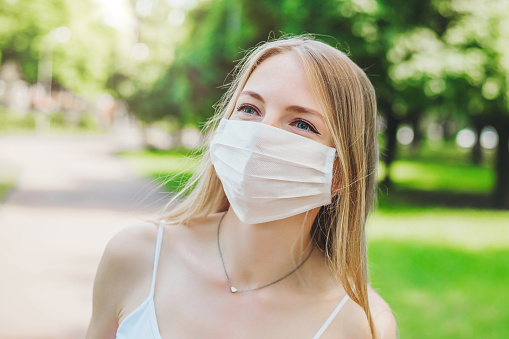 Portrait of a happy blonde student girl in a white medical mask smiling and walking in a park in the city, close up