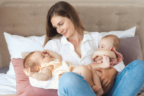 una giovane madre felice che tiene in braccio due bambine gemelle, seduta su un letto in camera da letto e sorridente alle sue due figlie bambine - twin newborn baby baby girls foto e immagini stock