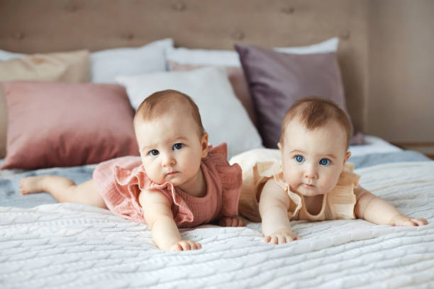 ritratto completo del corpo di due dolci sorelle gemelle di 6 mesi in abiti di cotone sdraiati su un comodo letto in una casa accogliente - twin newborn baby baby girls foto e immagini stock
