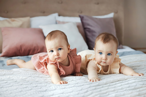Full body portrait of two sweet baby twins sisters 6 month in cotton dresses lying on comfortable bed at cozy home.