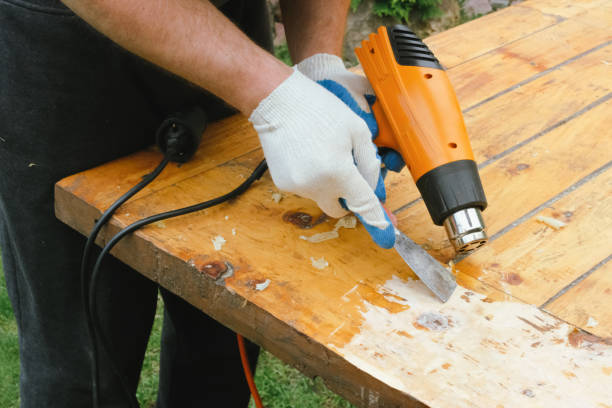 Man removing old varnish and paint from wood using scraper and heat gun. Restoration work of old wooden table outdoors. Furniture repair, reuse of old things concept.