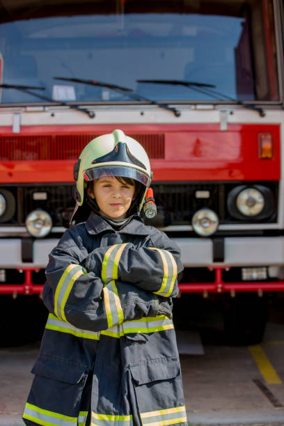 istock Child, cute boy, dressed in fire fighers cloths in a fire station with fire truck 1417612600