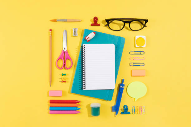 Yellow notebook, black pen, and white pencil on a light blue surface. A small metallic sharpener with shavings nearby. The background includes a decorative border with floral patterns. The composition suggests a workspace ready for writing or drawing. Light creates shadows, highlighting the objects' textures and colors.