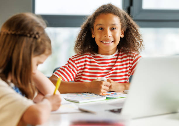 istock Cheerful  ethnic schoolgirl looking at camera  during   lesson with classmate 1417626750