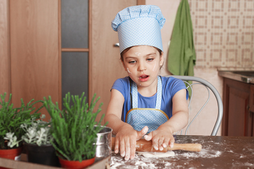happy 5 year old girl in blue dress and dressed as chef in kitchen rolling out the dough, lifestyle, selective focus