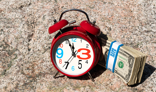 Red alarm clock and a stack of American bills on a stone