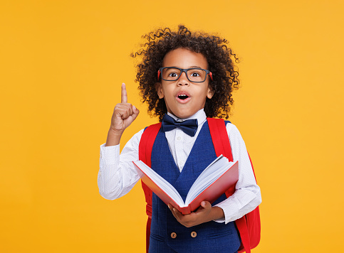Clever african american boy in glasses pointing up while reading textbook during school studies against yellow background