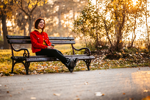 Beautiful young woman sitting on the park bench in the park during autumn, drinking coffee and listening to music via wireless headphones.