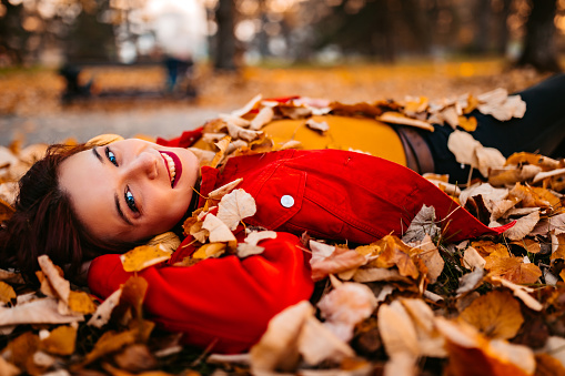 Beautiful young woman lying on autumn leaves in park and smiling.