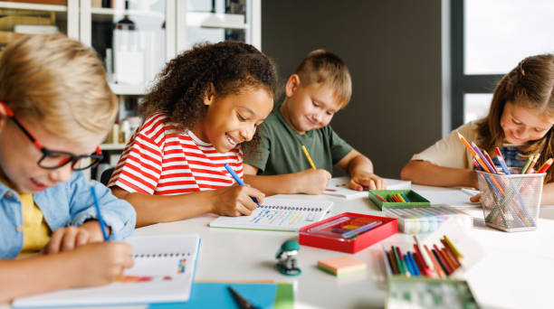 Cheerful ethnic girl solving test with classmates Positive ethnic student girl smiling and writing down data into notebook while sitting at table near classmates during test at school primary school student stock pictures, royalty-free photos & images